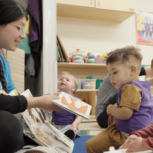 A caregiver sits on the floor reading to a group of toddlers, who are looking at picture books in a colorful classroom with toys and artwork on the walls.